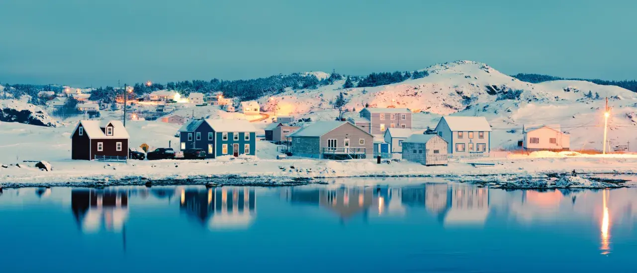 Snowy coastal village at dusk with colorful houses reflected in calm water.