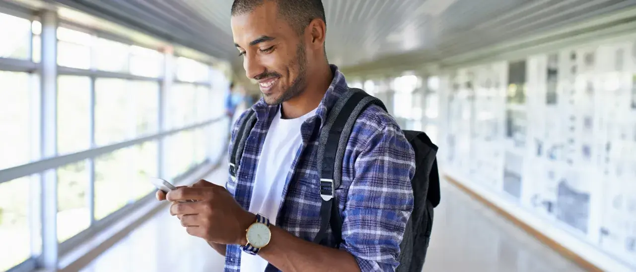 Man in plaid shirt with backpack looking at phone in a bright hallway.