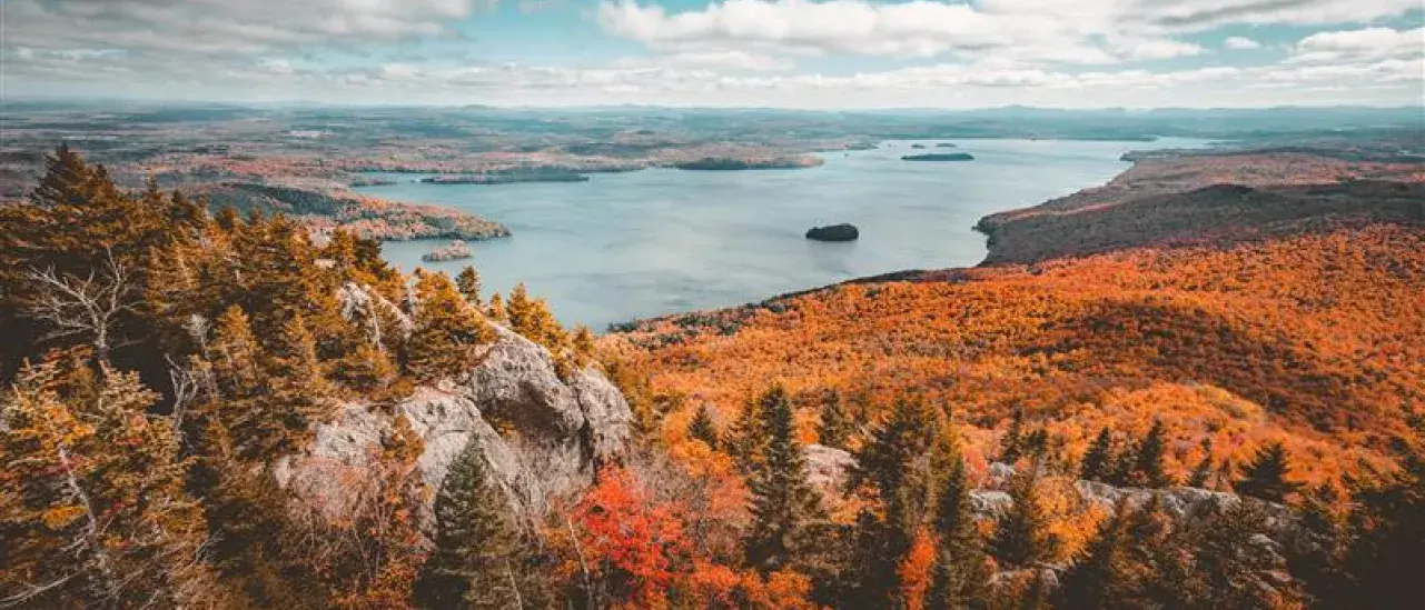 Scenic autumn landscape with a large lake surrounded by forested hills and mountains. Vibrant fall foliage in shades of orange, red, and yellow contrasts with the calm blue water, viewed from an elevated rocky vantage point under a partly cloudy sky.