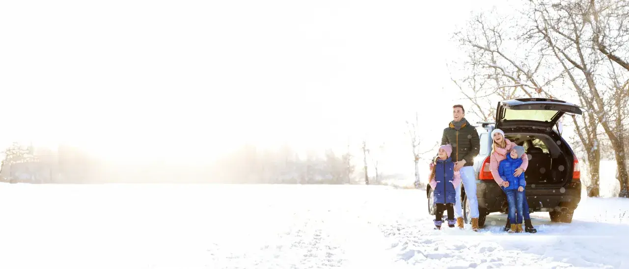 Family standing by an SUV with open trunk in a snowy field under bright sunlight.