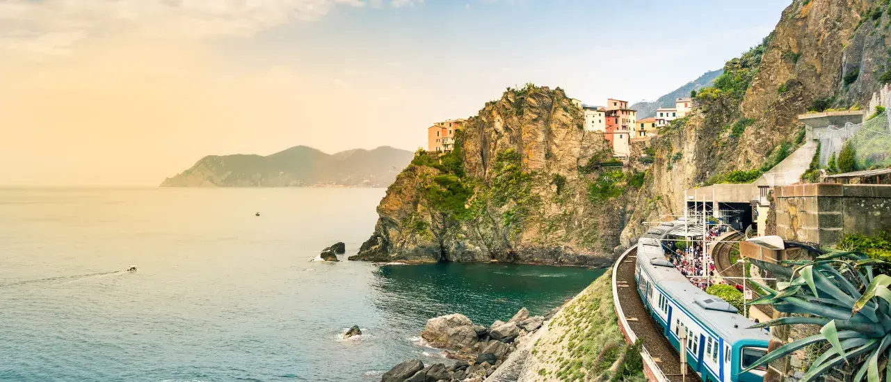 A train traveling along a coastal railway track on a steep rocky cliff overlooking the blue sea, with colorful buildings perched on the cliffside under a partly cloudy sky.