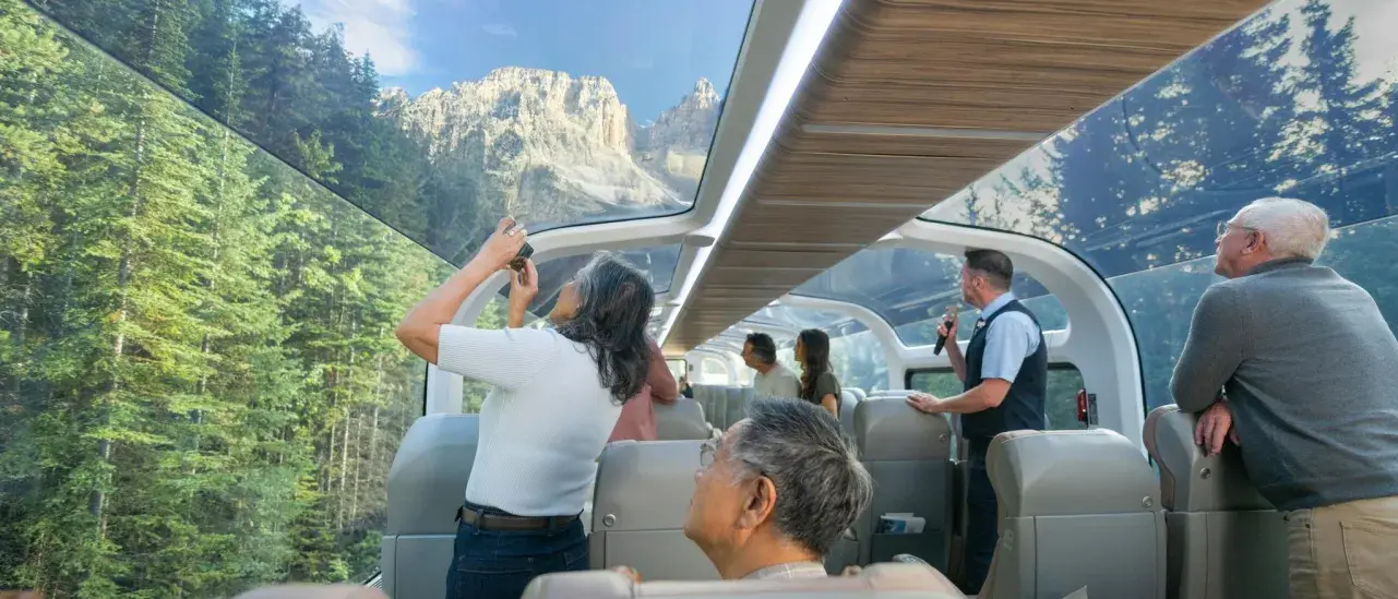 Scenic train interior with glass dome windows showing stunning mountain peaks and dense forest, passengers taking photos and enjoying panoramic views.
