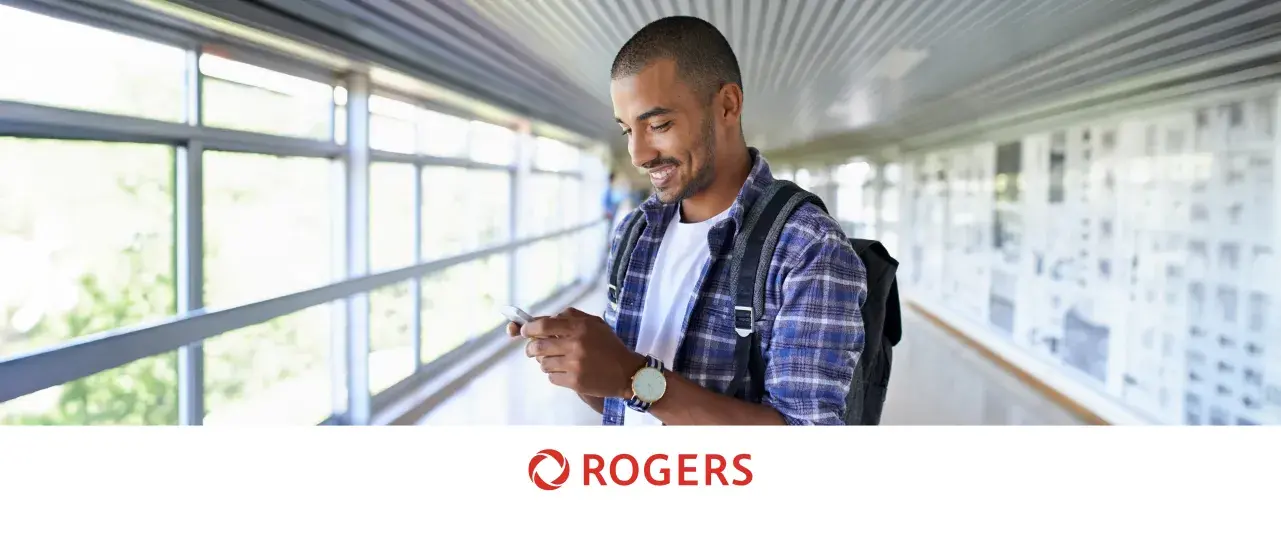 Man in plaid shirt with backpack looking at phone in a bright hallway.