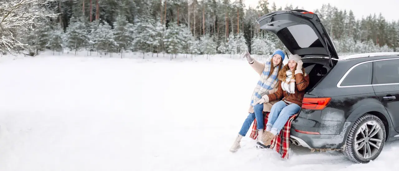 Two people sitting on the open trunk of a black SUV in a snowy landscape, with a forest in the background.