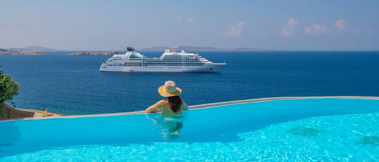 Person relaxing in an infinity pool overlooking a calm blue ocean with a large white cruise ship in the distance under a clear sky.
