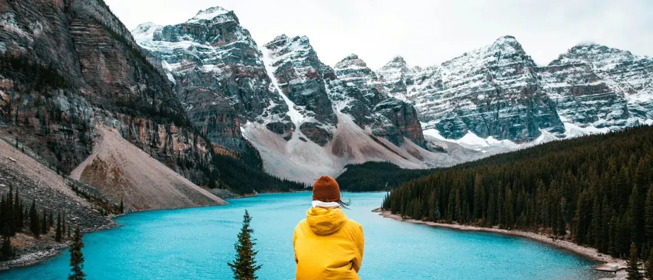 Person in a yellow jacket sitting on rocks overlooking a turquoise lake and snow-capped mountains.