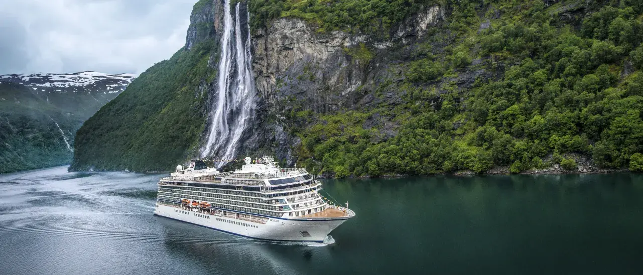 A large white cruise ship sails through a calm, deep‑green fjord surrounded by steep cliffs covered in lush greenery. A tall, narrow waterfall cascades down the cliffside near the ship under an overcast sky.