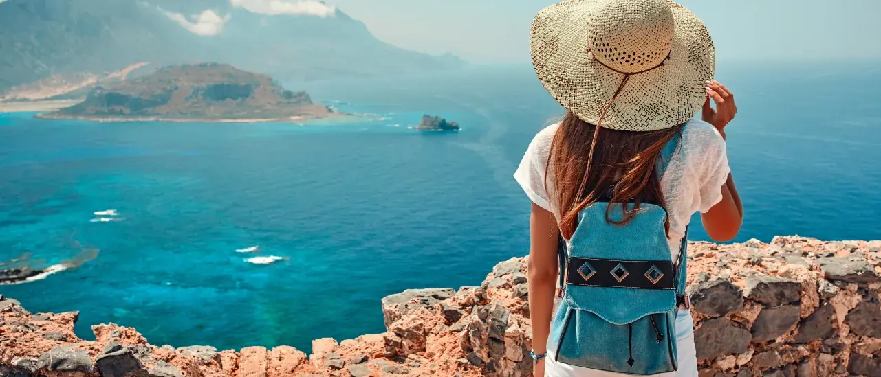 A person wearing a sunhat, white shirt, and a small blue backpack stands on a rocky viewpoint overlooking a bright blue sea with distant mountains and a small island in the background.