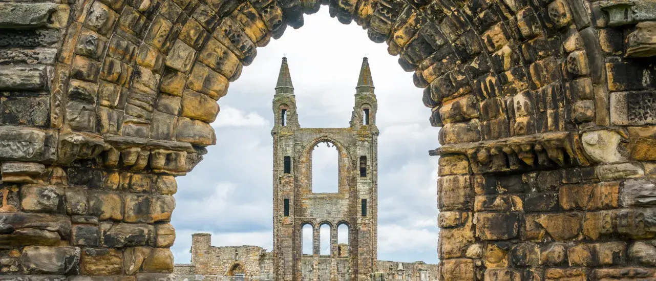 Stone archway framing the ruins of St Andrews Cathedral in St Andrews, Scotland, with a grassy courtyard and the cathedral’s central tower visible beyond.