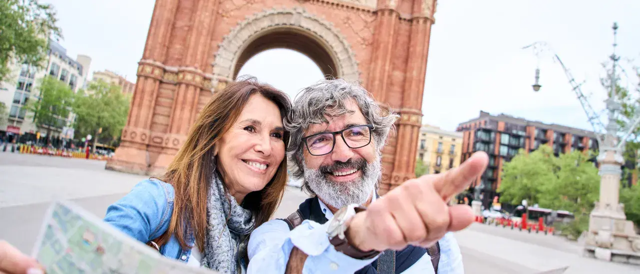 Two people stand in front of a large red-brick archway, holding a map as one of them points ahead. Buildings, trees, and a wide pedestrian walkway surround the landmark.