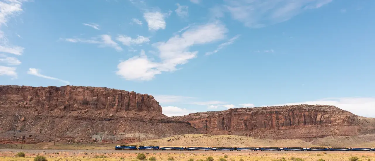 A long passenger train travels across a wide desert landscape with rugged red rock cliffs and a bright blue sky in the background.