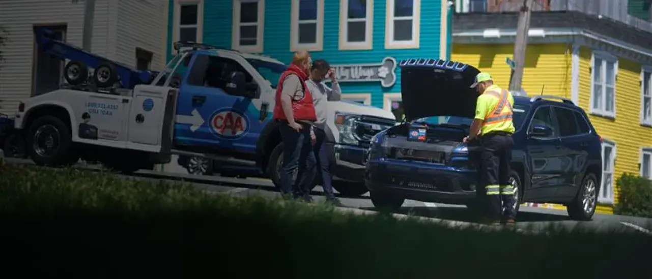 A CAA tow truck is parked on a street beside a dark SUV with its hood open, while a roadside assistance worker examines the engine and two people stand nearby talking. Colorful houses create a bright backdrop.