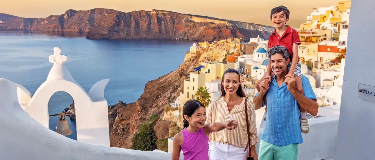 A family walks along a whitewashed pathway in a coastal village with blue-domed buildings, overlooking the sea and rugged cliffs at sunset.