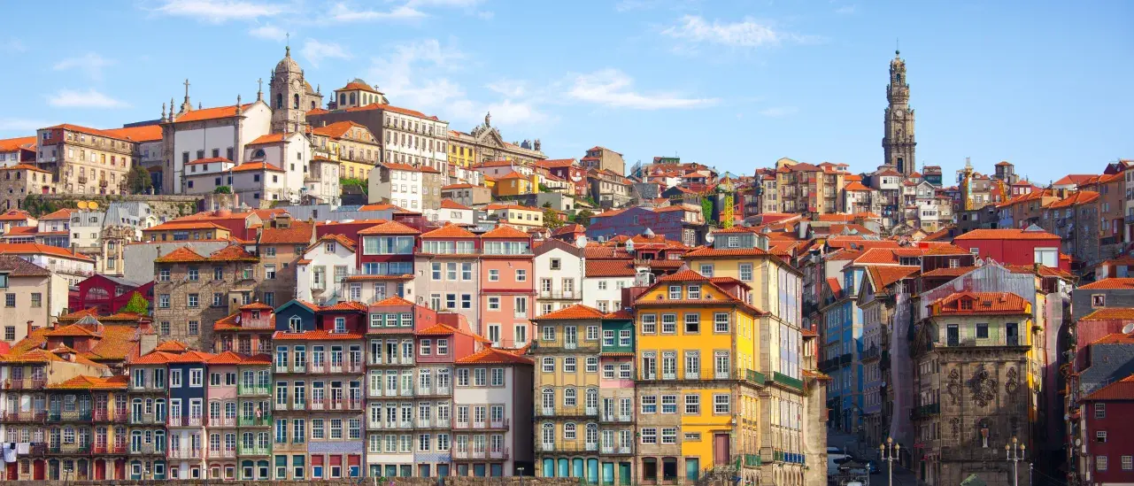 Colorful historic buildings with red-tiled roofs line the hillside along the Douro River in Porto, Portugal, with traditional boats moored on the water in the foreground and church towers rising above the city under a blue sky.