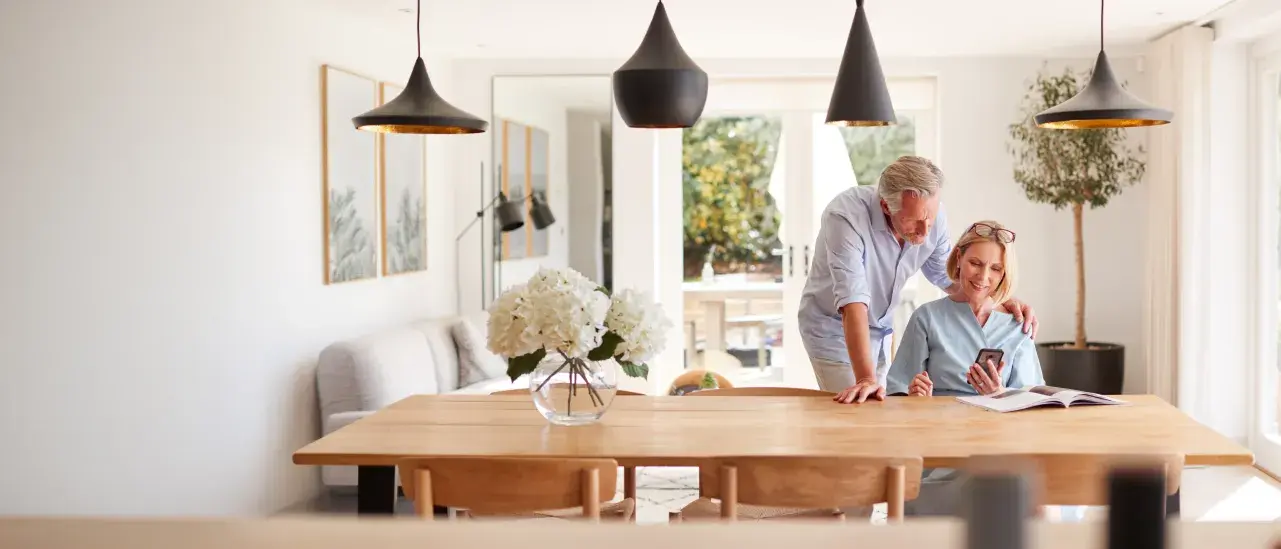 Two adults look at a smartphone together at a dining table in a bright, modern home.