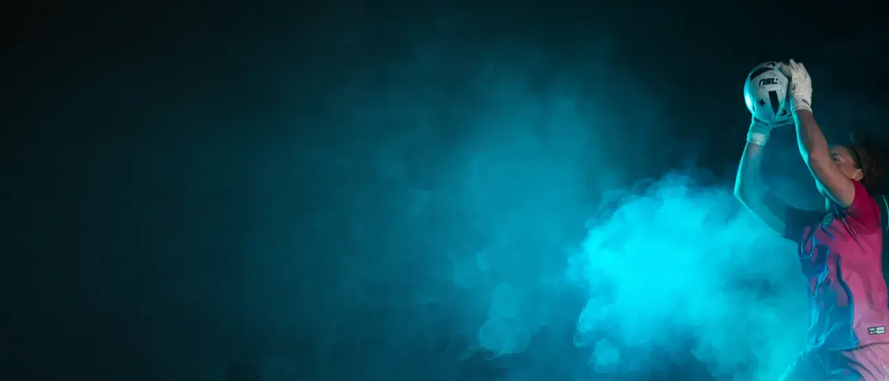 Athlete reaching up to catch a soccer ball in dramatic blue lighting against a dark background.