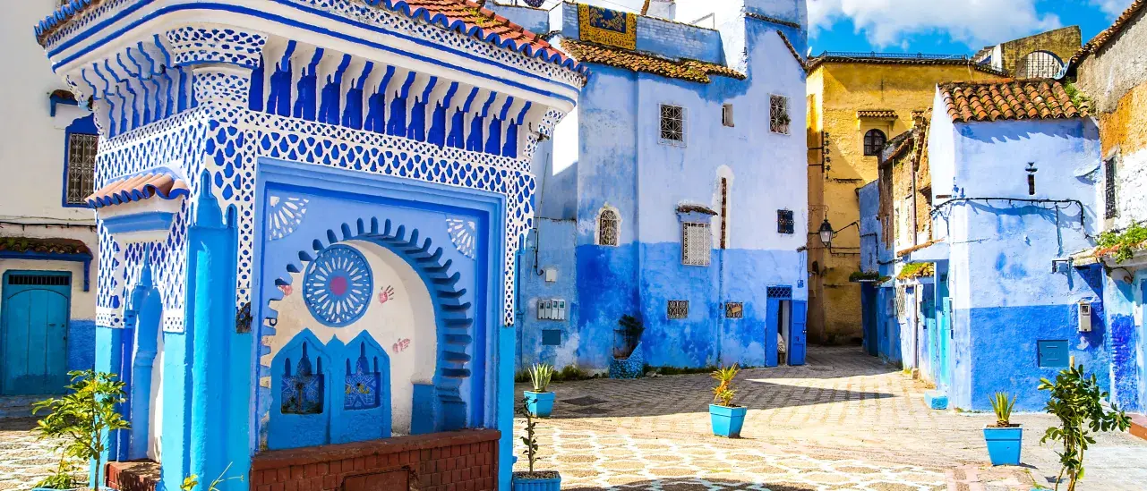 Blue and white buildings in a Moroccan street, featuring intricate architectural details, tiled rooftops, and a decorative fountain in the foreground under a bright blue sky.
