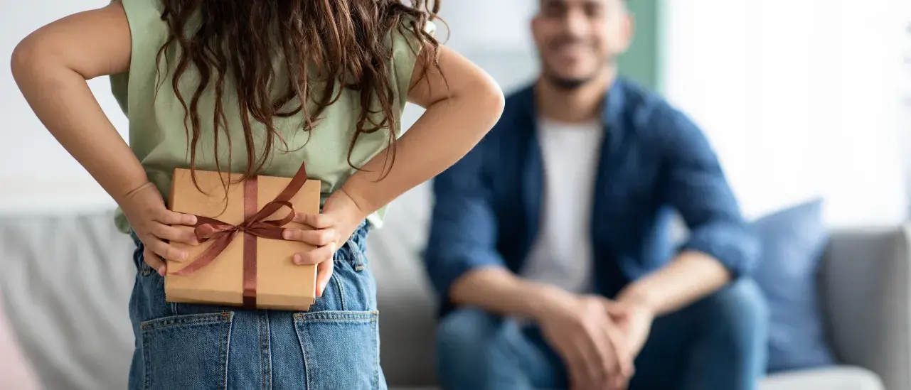 Child holding a wrapped gift behind their back while standing in front of an adult seated on a couch