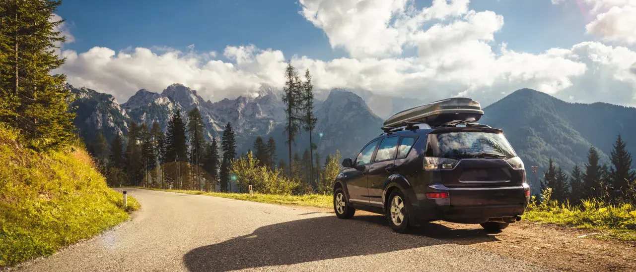  SUV parked on a winding mountain road with forested peaks and blue sky in the background.
