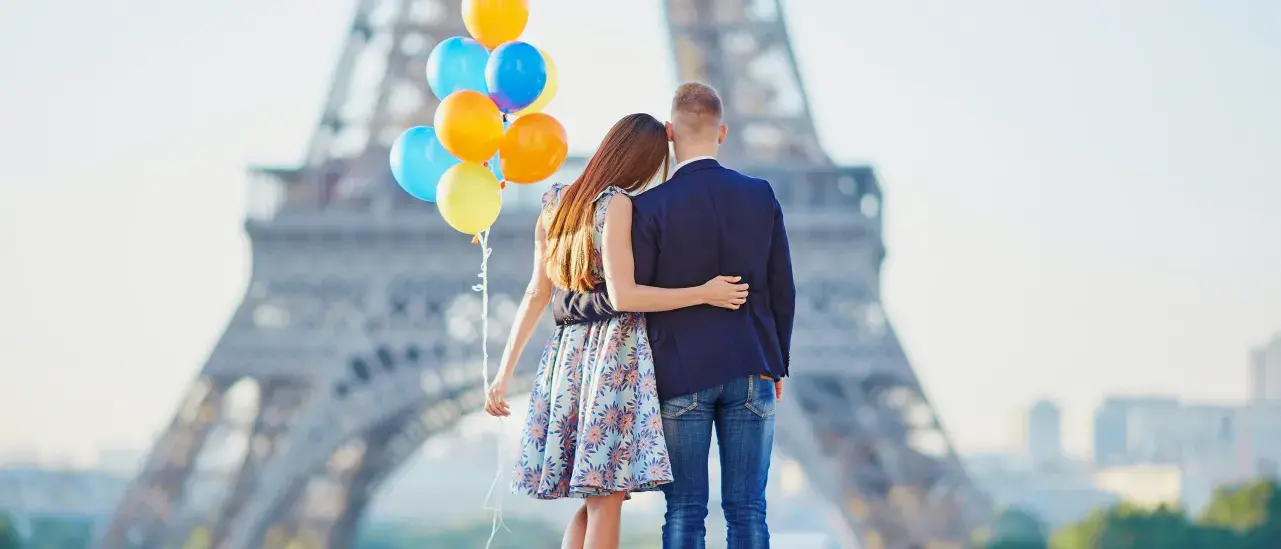 Two people walking away from the camera with arms around each other, holding a cluster of colorful balloons, with the Eiffel Tower blurred in the background on a bright day.