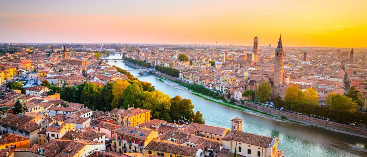 Aerial sunset view of Verona, Italy, with the Adige River, historic buildings, bridges, and church towers stretching across the cityscape.