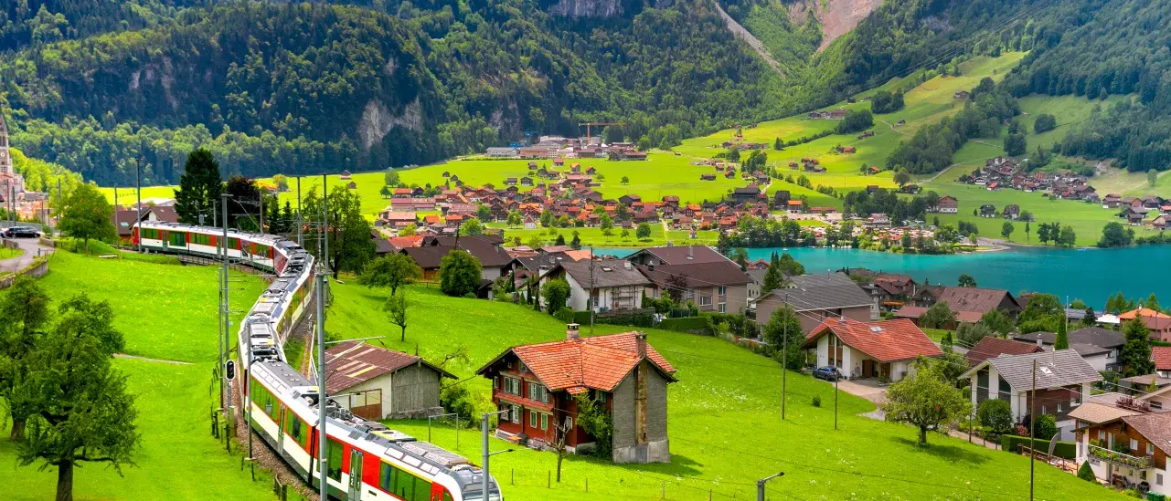 A passenger train winds through lush green countryside past scattered homes and farmland, with a turquoise lake, a village, and forested mountains rising in the background.