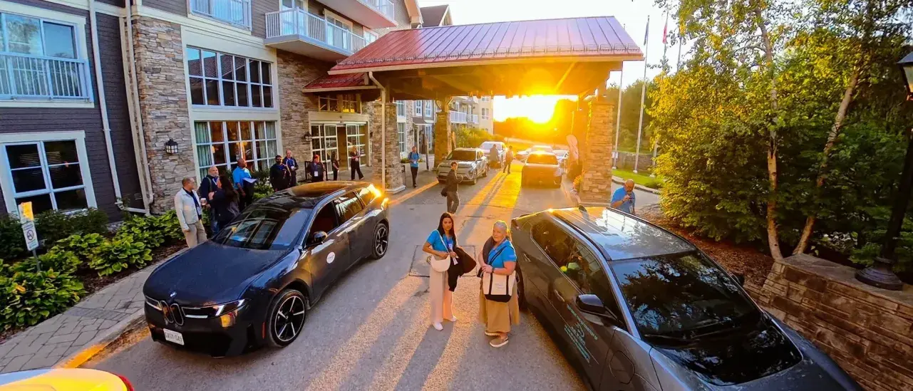 Vehicles arrive and guests gather outside the covered entrance of a multi-storey resort building at sunset, with warm sunlight casting long shadows across the driveway. Trees and landscaped grounds border the entrance area.