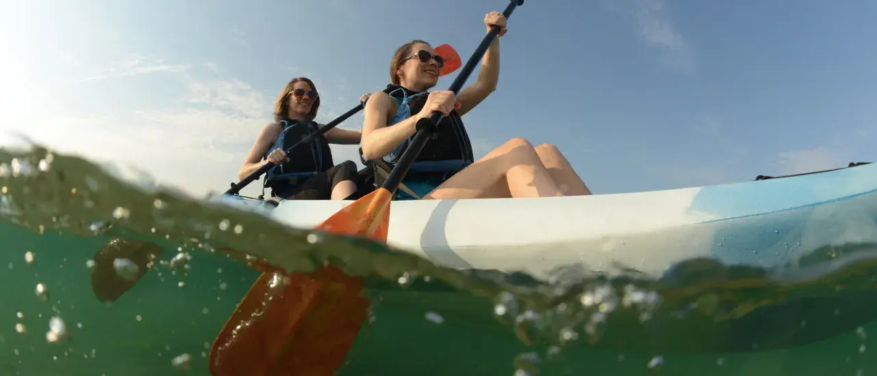 Two people kayaking on calm water under a blue sky, viewed from a split above-and-below-water perspective.
