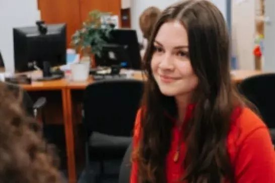 Female wearing a red quarter zip with long brown hair smiling in an office with computers in the background