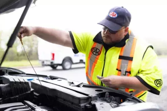 Roadside assistance worker in a bright saftey vest checking under the hood of a car on a highway shoulder with a white truck in the background