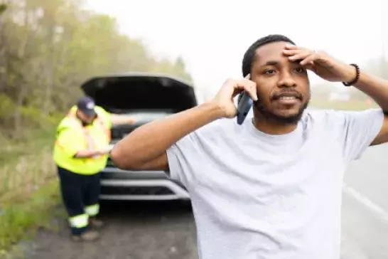 Man wearing white tshirt on the shoulder of a highway looking a bit distressed while on the phone with his hand on his forehead. Roadside assistance worker wearing neon safety gear under the roof of car in the background