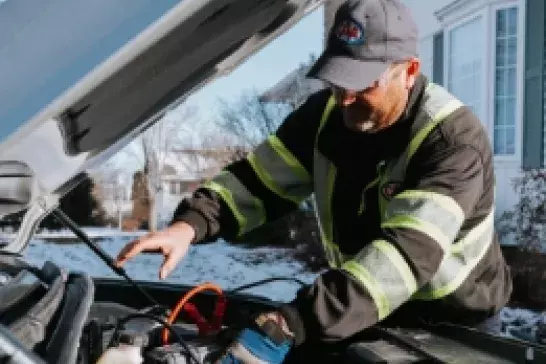 Roadside assistance worker wearing a ball cap and safety gear working under the hood of a vehicle 