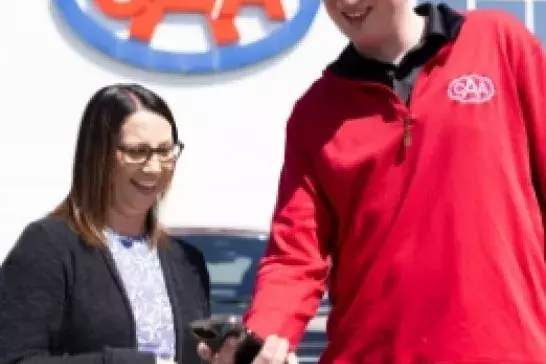 tall male CAA worker in a red shirt showing a shorter female wearing a black sweater and glasses something on his phone with the CAA logo on the side of a building in the background