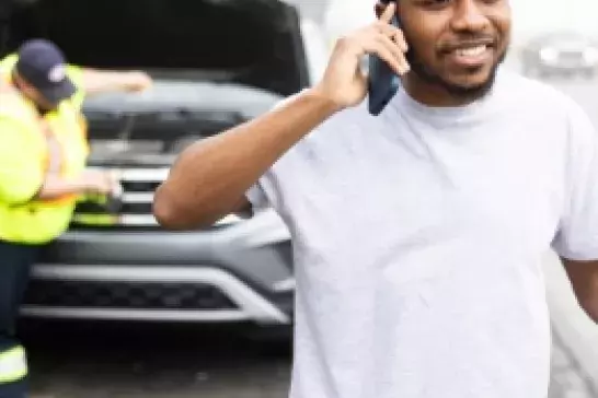 african-american male on the shoulder of a highway wearing a white tshirt holding a phone to his ear with a roadside technician in the background attending to a car with the hood propped open.