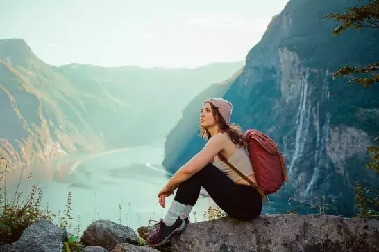 A person wearing a beanie, tank top, leggings, and hiking shoes sits on a rock with a backpack. They are in a mountainous area with a river and waterfall in the background, looking contemplatively into the distance.