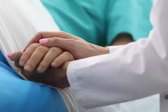close up shot of a doctors hand with pink nailpolish holding a patients hand while they are laying in a bed with a blue blanket