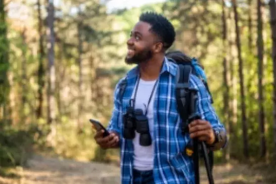 Male holding a phone and walking stick wearing a blue plaid shirt over a white shirt, a backpack and binoculars slung over his neck with trees in the background