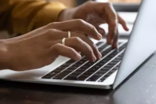 Two hands typing on a laptop keyboard on a brown tabletop. One hand has two silver rings on two fingers