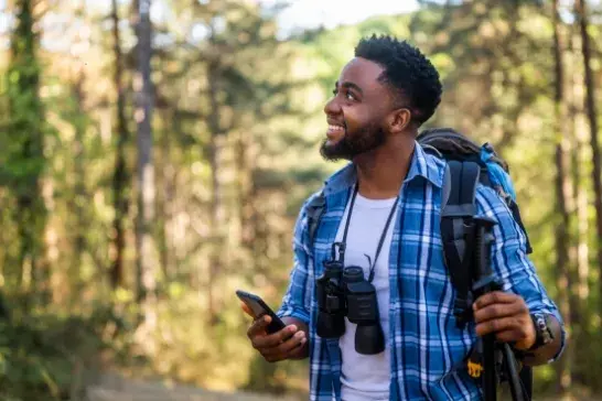 Male holding a phone anda. walking stick wearing a blue plaid shirt over a white shirt, a backpack and binoculars slung over his neck with trees in the background