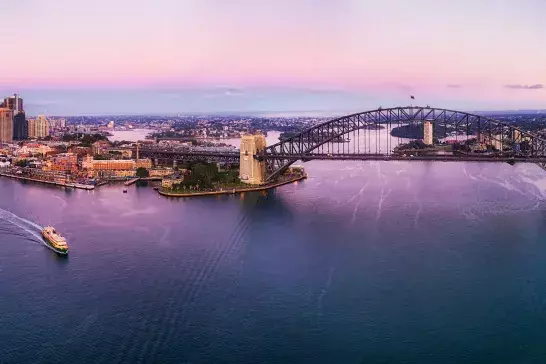 A panoramic aerial view of Sydney Harbour at sunset, featuring the Sydney Opera House on the left and the Sydney Harbour Bridge in the center. The city skyline is visible with numerous buildings, and the sky is painted with pink and purple hues.