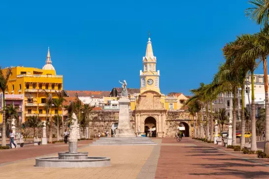 A city plaza featuring a clock tower, several statues, palm trees, and surrounding buildings under a clear blue sky.