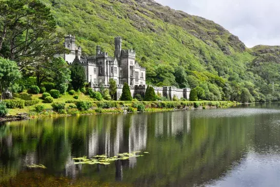 A castle with towers and turrets is situated beside a lake, surrounded by lush greenery and hills.