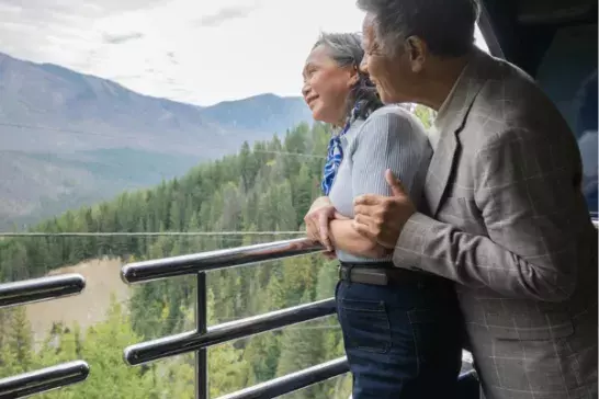 Two people stand on a balcony or observation deck, overlooking a scenic view of mountains and forests. The individuals are facing away from the camera, with one person holding the other. The background features lush green trees and distant mountains under a cloudy sky.