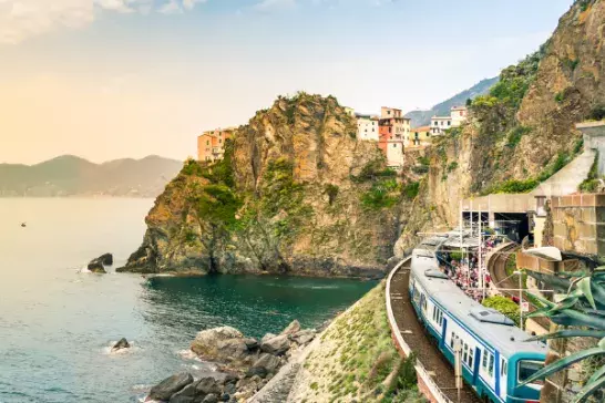 A train traveling along a coastal railway track that curves around a rocky, green-covered cliff. Buildings sit atop the cliff, and the calm sea below reflects the warm, golden light of the setting or rising sun under a partly cloudy sky.