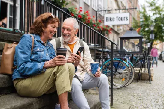 Two people sitting on outdoor steps in an urban area, holding cups and looking at a smartphone. A "Bike Tours" sign is visible in the background, along with bicycles, buildings, and cobblestone pavement.