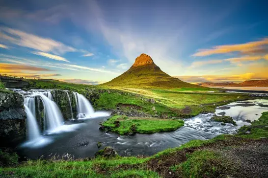 A cone-shaped mountain with a grassy foreground and a waterfall cascading into a river on the left. The sky is clear with some clouds illuminated by the setting or rising sun.