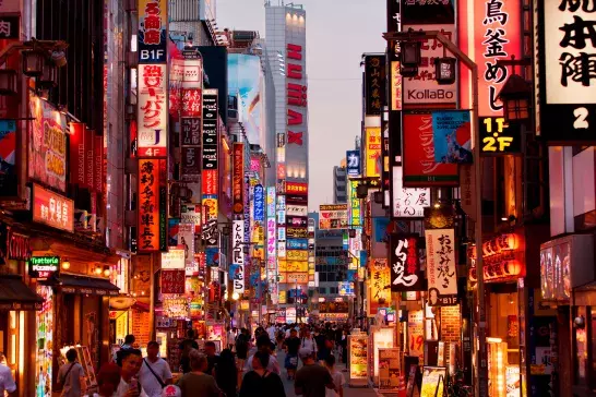 A bustling street in Japan filled with numerous brightly lit signs and advertisements in Japanese. The street is crowded with people walking, and the buildings are adorned with colorful neon lights and billboards.