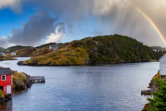 A picturesque coastal scene with a small inlet of water surrounded by grassy hills and scattered houses. A red wooden house is near the water's edge on the left, while white wooden houses are on the right. A rainbow arches across the sky above the hills in the background, under a partly cloudy sky.