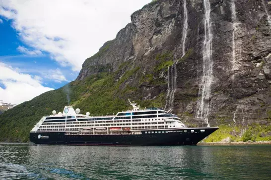 A large cruise ship sailing near a steep, rocky cliff with multiple waterfalls cascading down the cliffside. The water is calm, and the sky is partly cloudy with patches of blue. The ship has several decks and various structures on top, including domes and antennas.