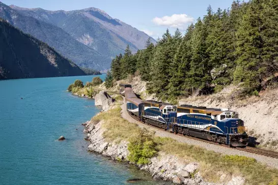 The Rocky Mountaineer train traveling along a scenic route beside a turquoise lake, surrounded by lush green trees and mountains in the background. The train is blue with white and gold accents, and the words "ROCKY MOUNTAINEER" are visible on the side of the lead locomotive.